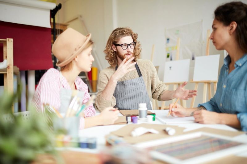 Young students in workwear sitting by table in studio and discussing creative ideas at meeting