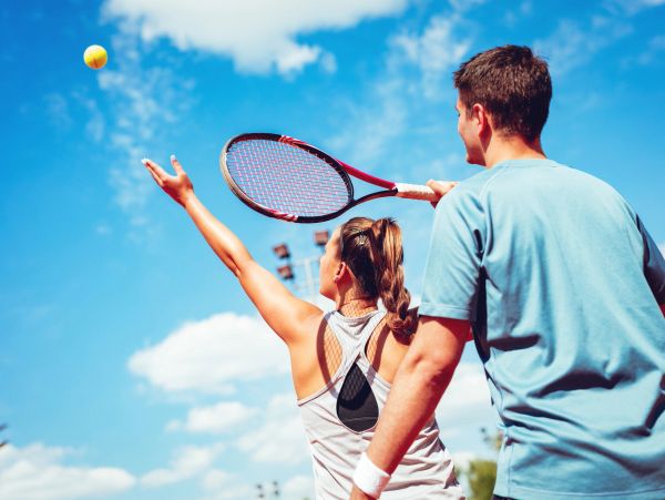 Woman serving a tennis ball with a man watching.