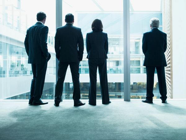 Four business professionals stand facing a large window in an office building.