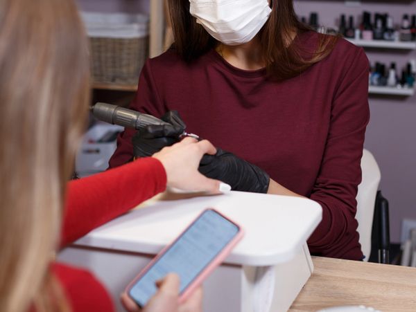 Manicurist wearing mask and gloves working on client's nails in a salon.