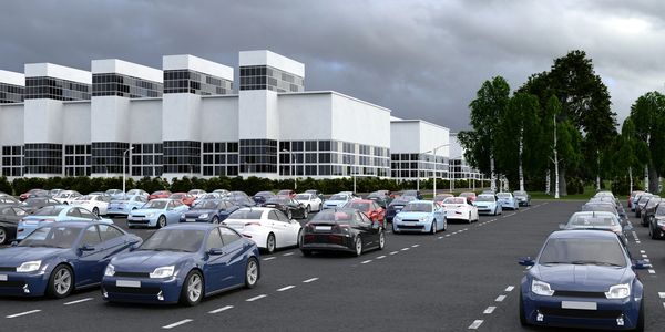 A large parking lot filled with cars near modern white buildings under a cloudy sky.