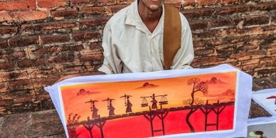 A man displays a vibrant painting of people crossing a bridge at sunset.
