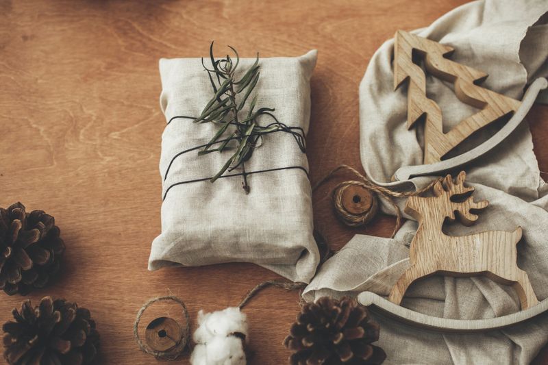 Stylish christmas rustic gift wrapped in linen fabric with green branch on wooden table with pine cones, tree,reindeer, twine, cotton. Simple eco presents plastic free. Zero waste  holidays