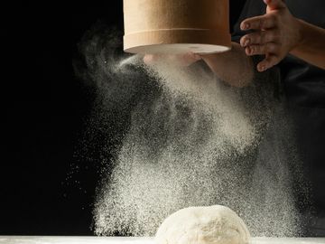 Hands sifting flour over dough on a dark background.