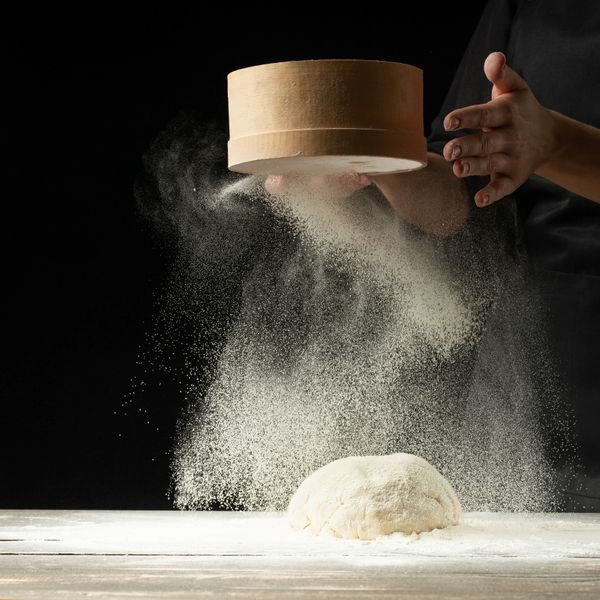Hands sifting flour over dough on a wooden surface against a black background.
