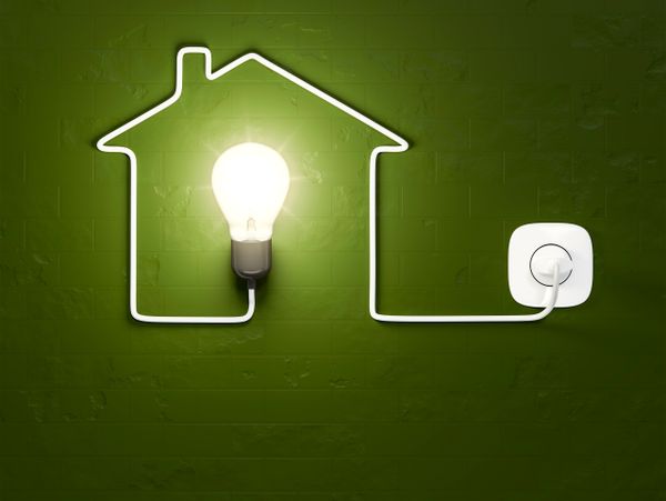 Light bulb shaped as a house connected to a power socket on green wall.