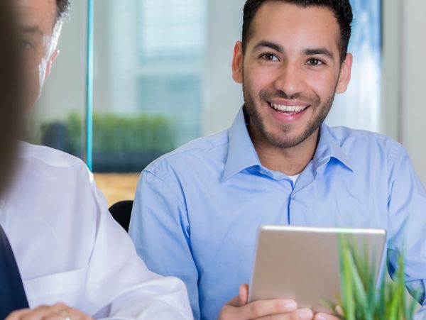 A smiling young man in a blue shirt holding a tablet in a meeting.