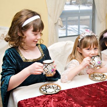Two girls in princess dresses having tea indoors by a window.