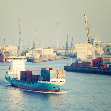 Cargo ship carrying containers sailing through a busy port with cranes and docks.