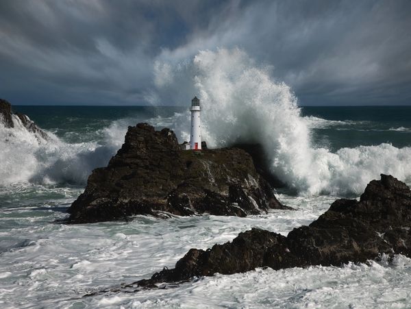 Powerful ocean waves crash around a lighthouse on rocky cliffs under a cloudy sky.