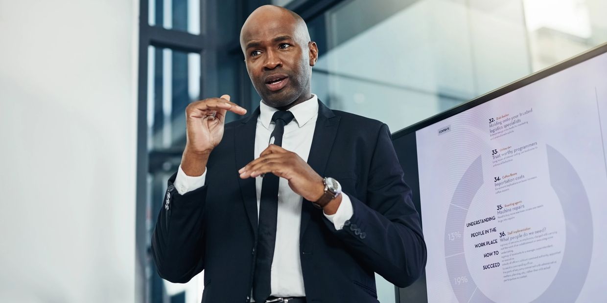 A businessman gives a detailed presentation with a chart on a screen behind him.
