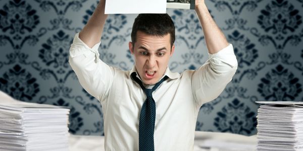 Stressed man angrily holding papers above his head at a cluttered desk.