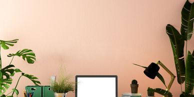 A cozy workspace with wooden furniture, plants, and a laptop against a peach wall.