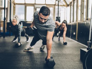 Three people working out with dumbbells in a gym, balancing in plank position.