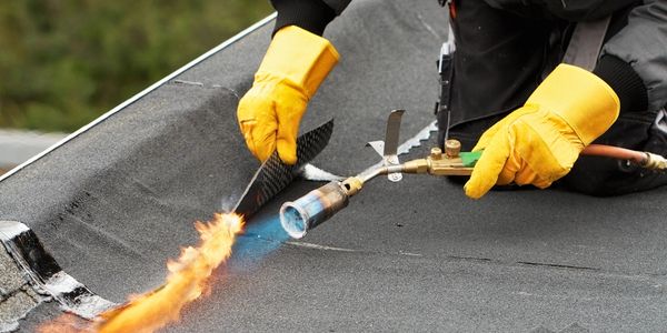 Worker using a blowtorch to seal roofing material while wearing protective gloves.