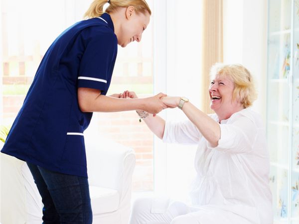 Caregiver helping elderly woman sit on an exercise ball, both smiling warmly.
