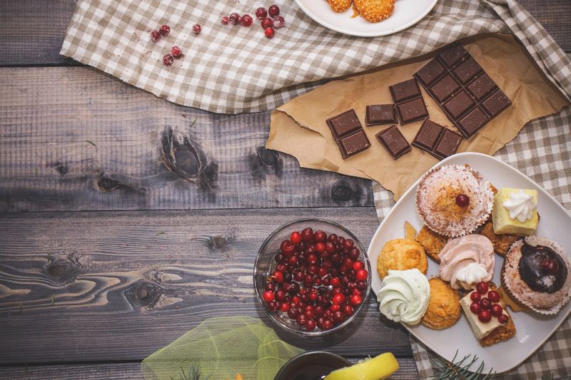 Close-up of sweets on a white plate: coconut pechente, pastila, meringue, cream roses, razat Turkish delight, near a broken chocolate bar on craft paper, in a bowl sesame cookie in a bowl on a linen flax napkin, fresh red cranberries, a glass with a dark drink decorated with a lemon slice little spruce branches. Festive pastry composition.