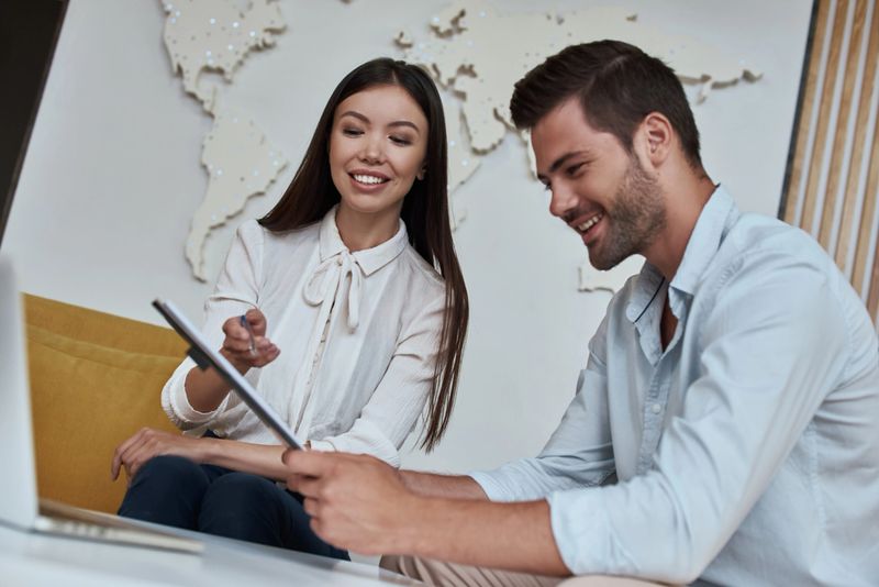 Young man reviewing contract for a trip with agent sitting at the travel agency office with world map on the background. Young woman offering vacation options for male tourist.