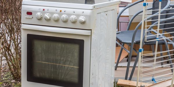 Discarded white electric stove outdoors with surrounding furniture and debris.