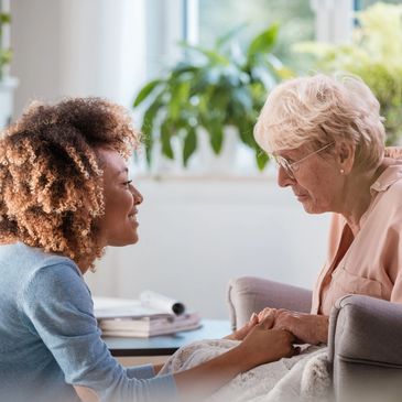 A young woman compassionately holds hands with an elderly woman in a cozy living room.
