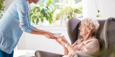 A caregiver offers a cup to an elderly woman sitting in a chair.