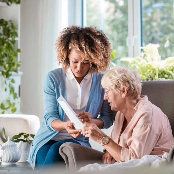 A younger woman assists an elderly woman with a document in a cozy living room.