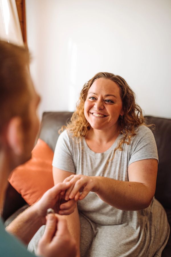 A woman smiling as a man proposes with a ring at home.