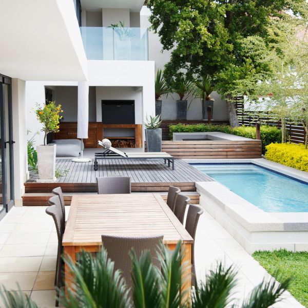 Modern backyard with wooden dining set, lounge chair, and pool surrounded by greenery.
