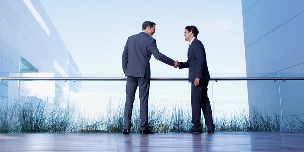 Two businessmen shaking hands outdoors between modern buildings.