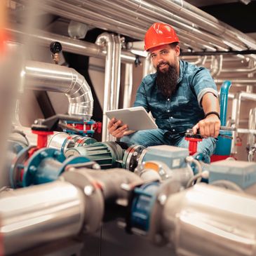 Engineer with red helmet inspecting industrial pipes and machinery holding a tablet.