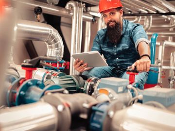 A technician in a hard hat working with industrial pipes and a tablet.