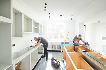 Two men installing kitchen cabinets in a bright room.