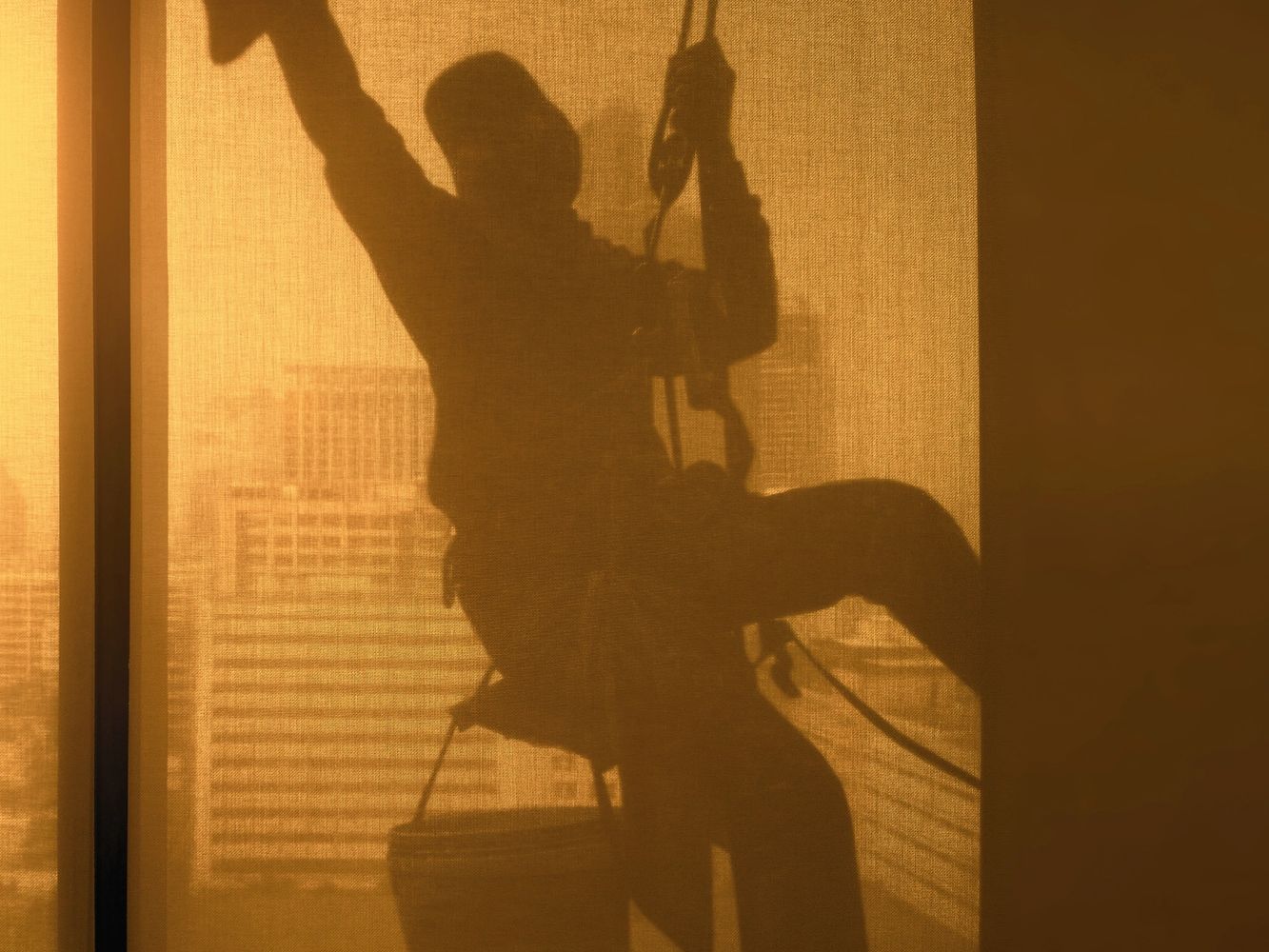 Silhouette of a window cleaner suspended outside a building at sunset.