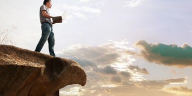 Man standing on eagle-shaped rock with laptop at sunset.