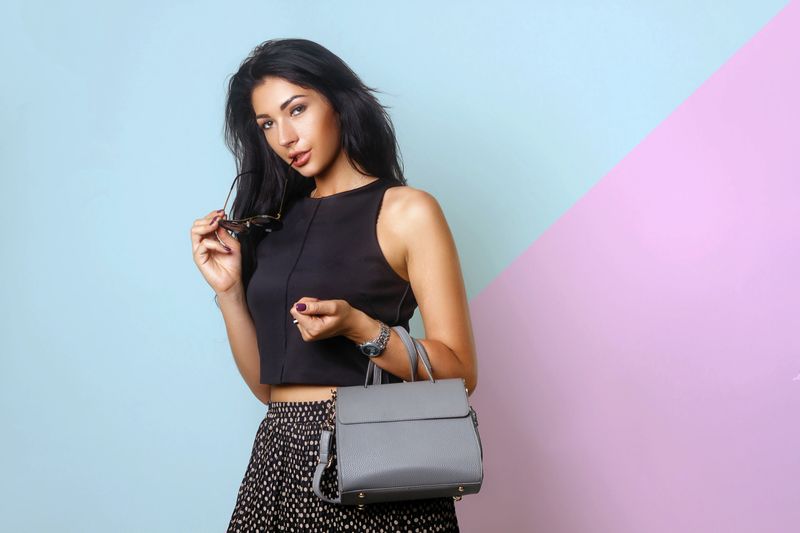 Portrait of a beautiful girl posing in studio in black blouse, skirt and fashion sunglasses, holding handbag. The concept of stylish and sexy women, perfect skin and body on aqua pink background