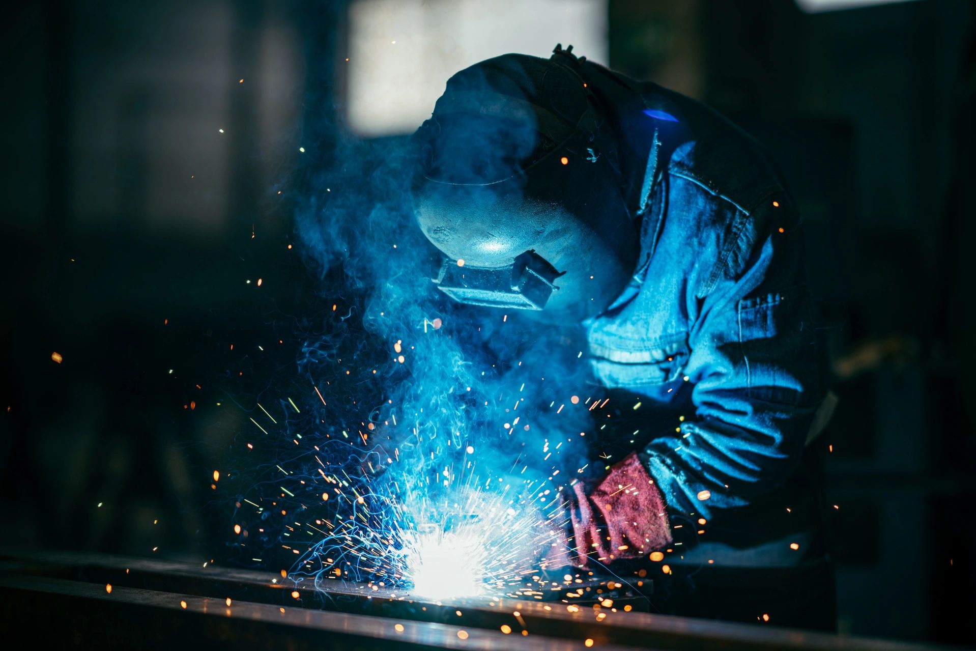 Man wearing welding helmet, welding in machine shop. Bright sparks and blue haze flying in the air.