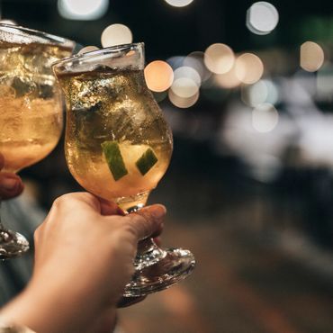 Two hands holding glasses of iced cocktails with lime, blurred lights in background.