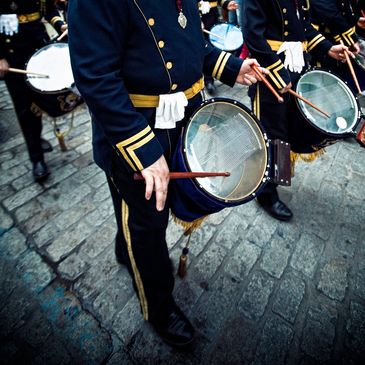 Drummers in uniform playing drums during a parade on a cobblestone street.