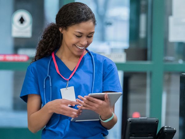 Smiling nurse in blue scrubs using a tablet in a hospital.