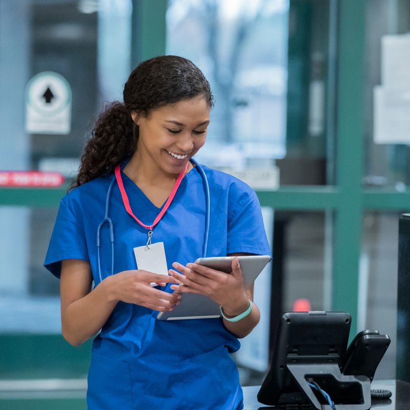 Young adult mixed race woman checks a patient's chart on a digital tablet while standing at a nurses station in hospital emergency room.