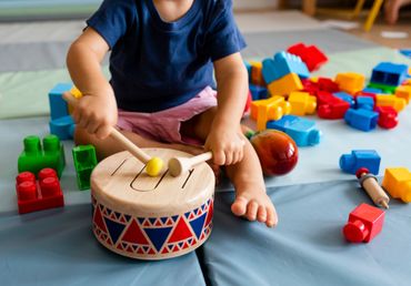 Toddler playing a wooden drum surrounded by colorful building blocks.