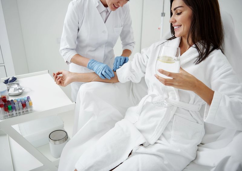 Cropped portrait of beautiful woman in white bathrobe sitting in armchair and receiving IV infusion. She is holding glass of beverage with lemon and smiling