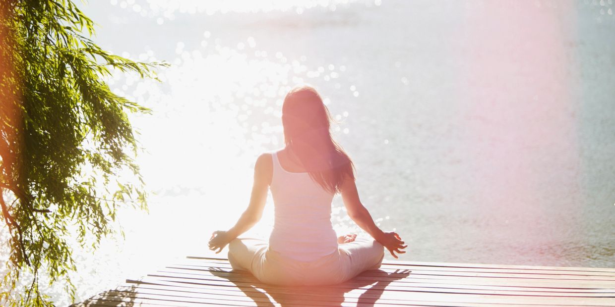 Person meditating by water in sunlight.