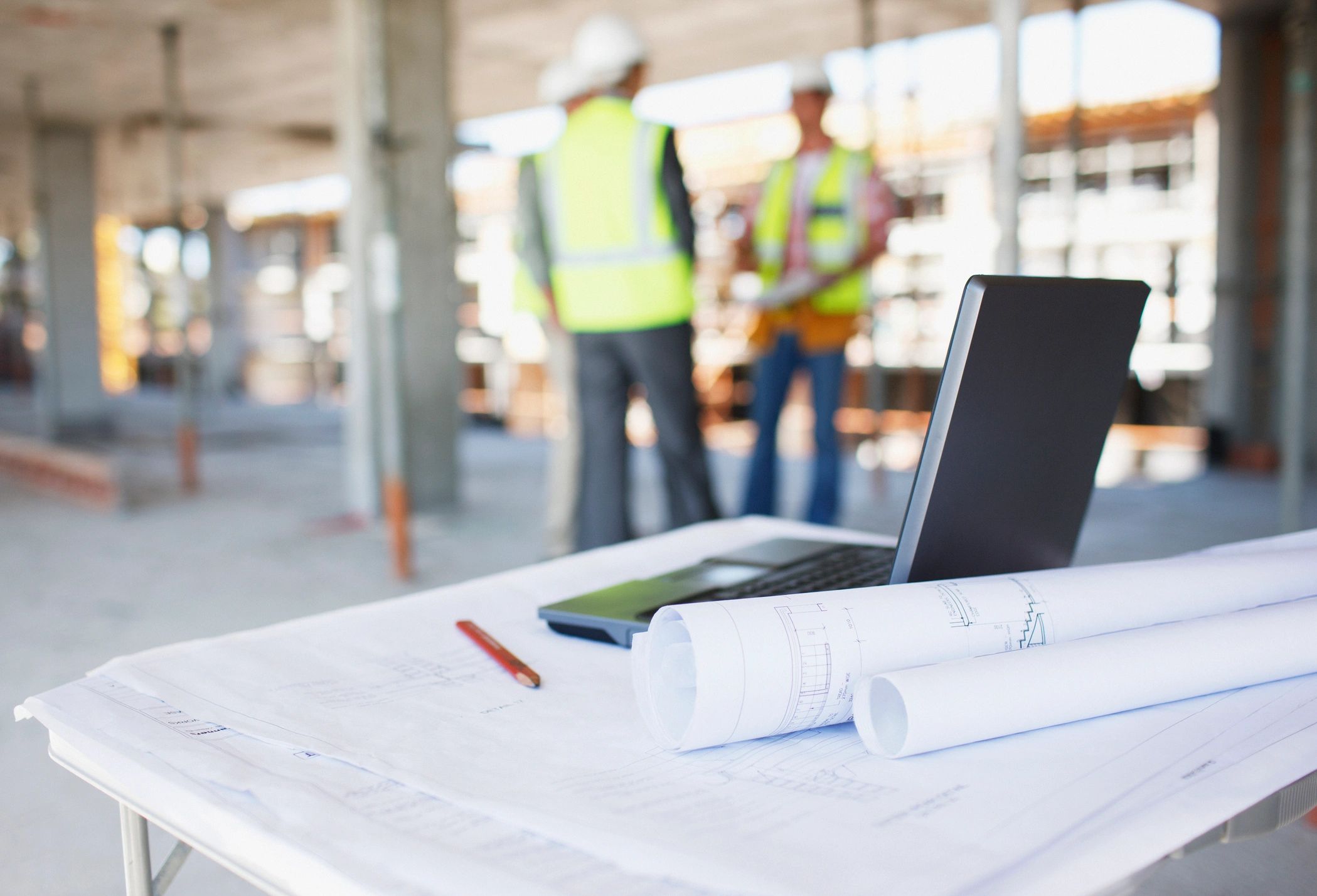 Blueprints and laptop on a table at a construction site with workers in the background.