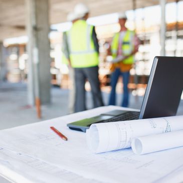 Blueprints, a laptop, and a tablet on a table at a construction site with workers in safety vests.