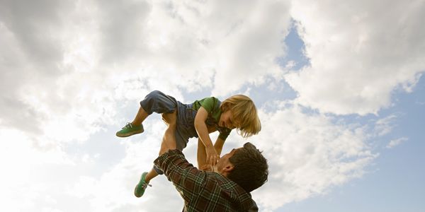 A man lifts a child playfully against a cloudy sky backdrop.