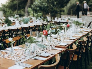 Elegant outdoor dining setup with wooden tables, floral centerpieces, and greenery.