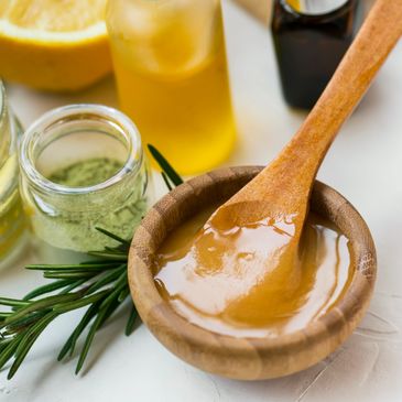 Wooden bowl with honey and a wooden spoon surrounded by herbs and lemon.