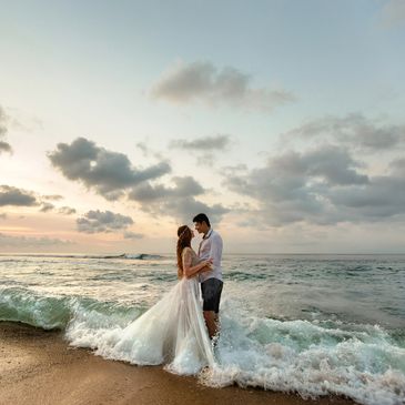 Bride and groom stand together in the surf, holding hands with waves gently around their feet.
