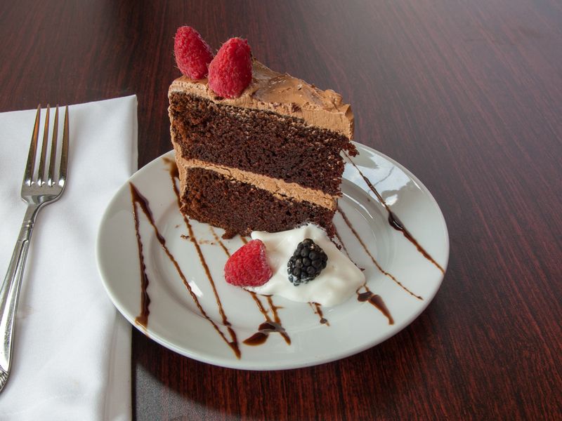 a beautiful slice of chocolate cake with chocolate frosting with a chocolate syrup drizzle complete with a dollop of whip cream and fresh raspberries and blackberries